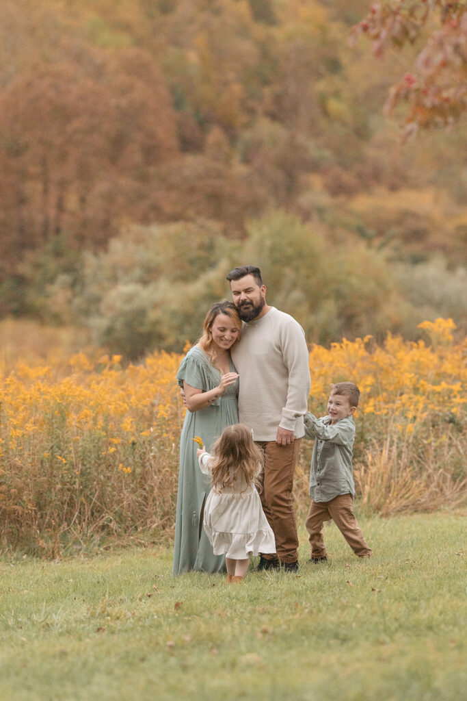 A mom and dad stand in a field by some wildflowers as their two young children play around them