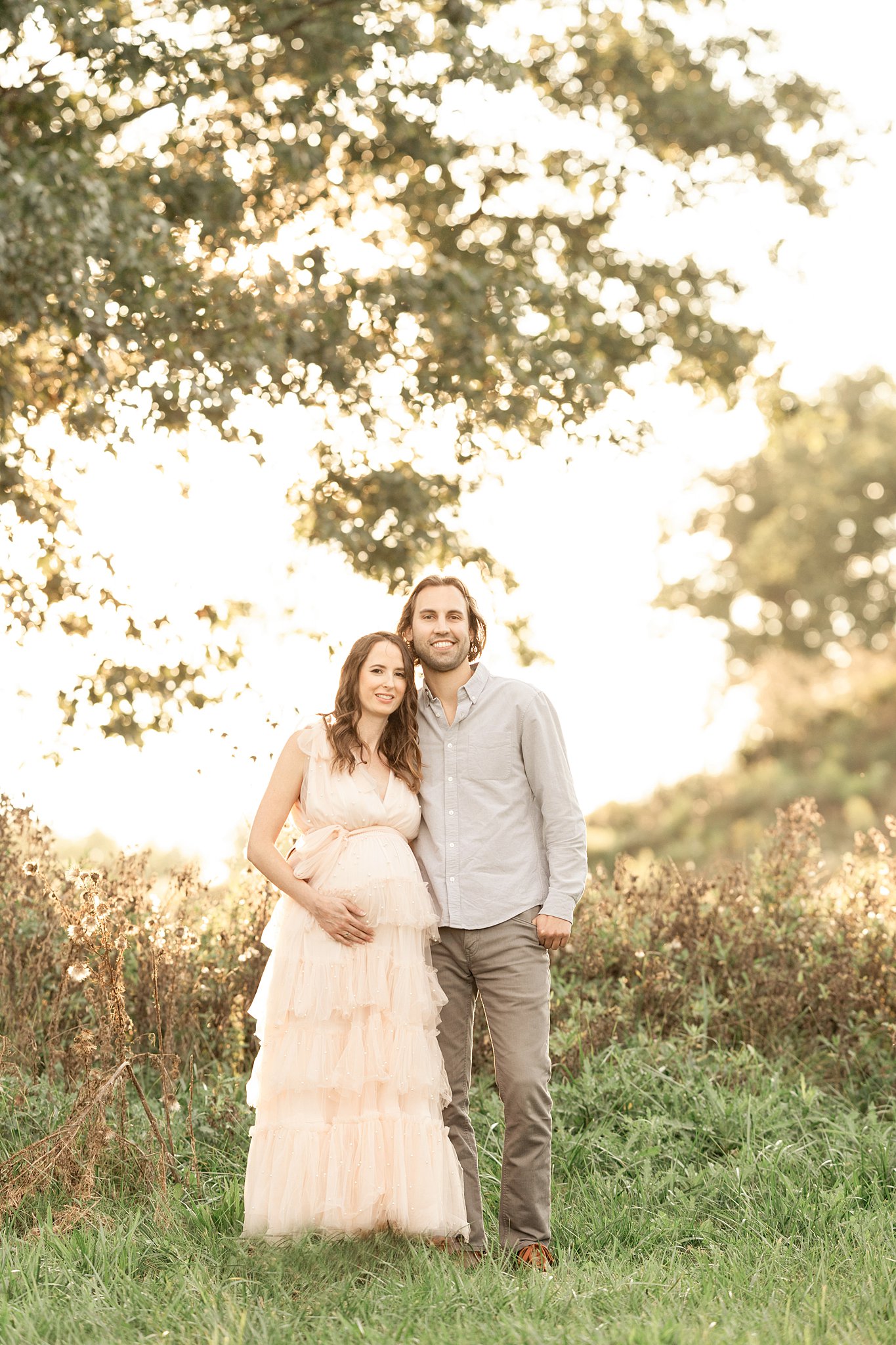 A mom to be in a white maternity gown leans into her partner as they stand in a field of grass