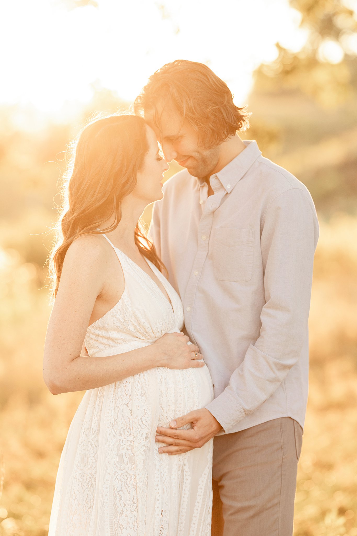 A mom to be nuzzles with her partner as they hold the bump and stand in a field at sunset
