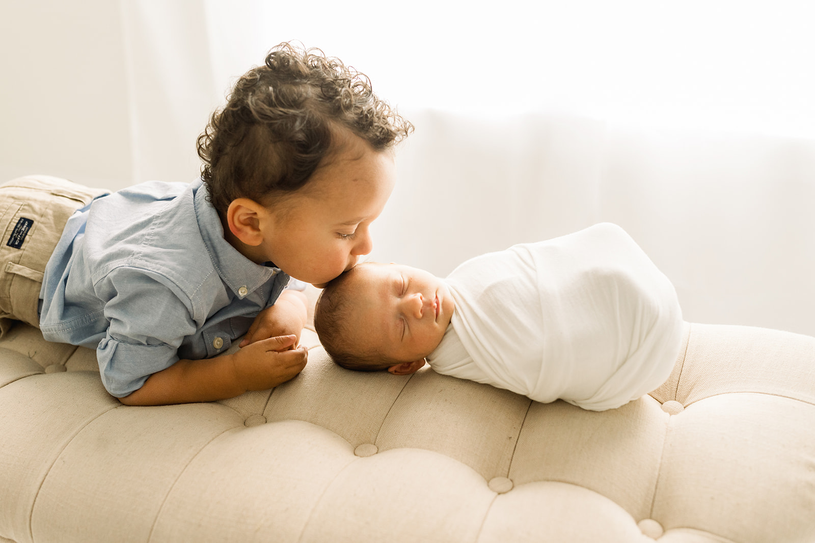 A young boy in a blue shirt kisses the head of his sleeping newborn baby sibling on a tan couch by a window
