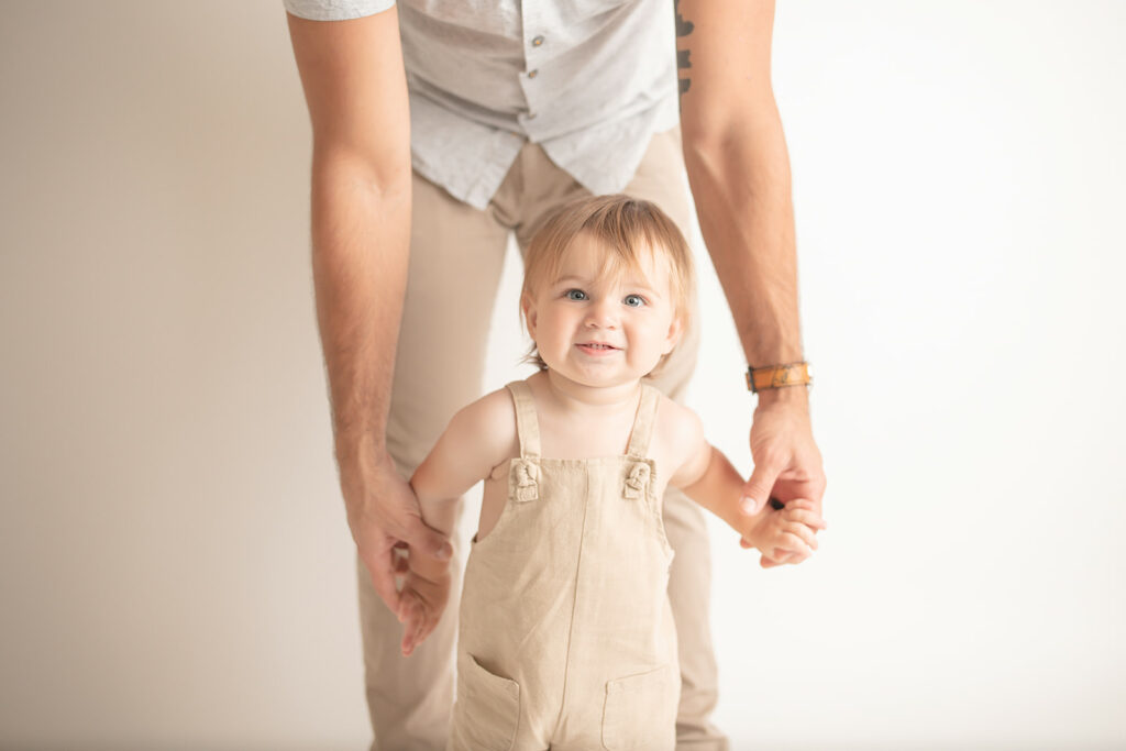 A toddler boy walks through a studio holding dad's hands in tan overalls while smiling thanks to Pittsburgh Pediatric Dentistry
