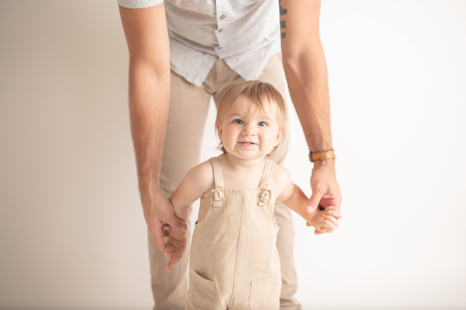 A toddler boy walks through a studio holding dad's hands in tan overalls while smiling thanks to Pittsburgh Pediatric Dentistry