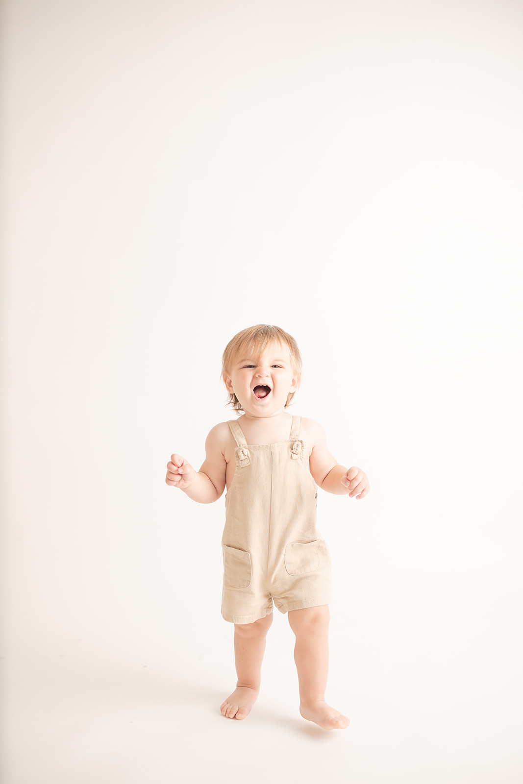 A young toddler boy makes silly faces while walking through a studio in tan overalls