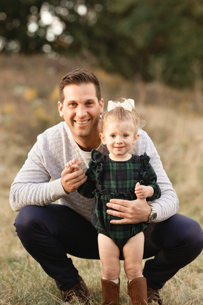 A father in a grey sweater squats with his toddler daughter in a field of grass in a green plaid dress
