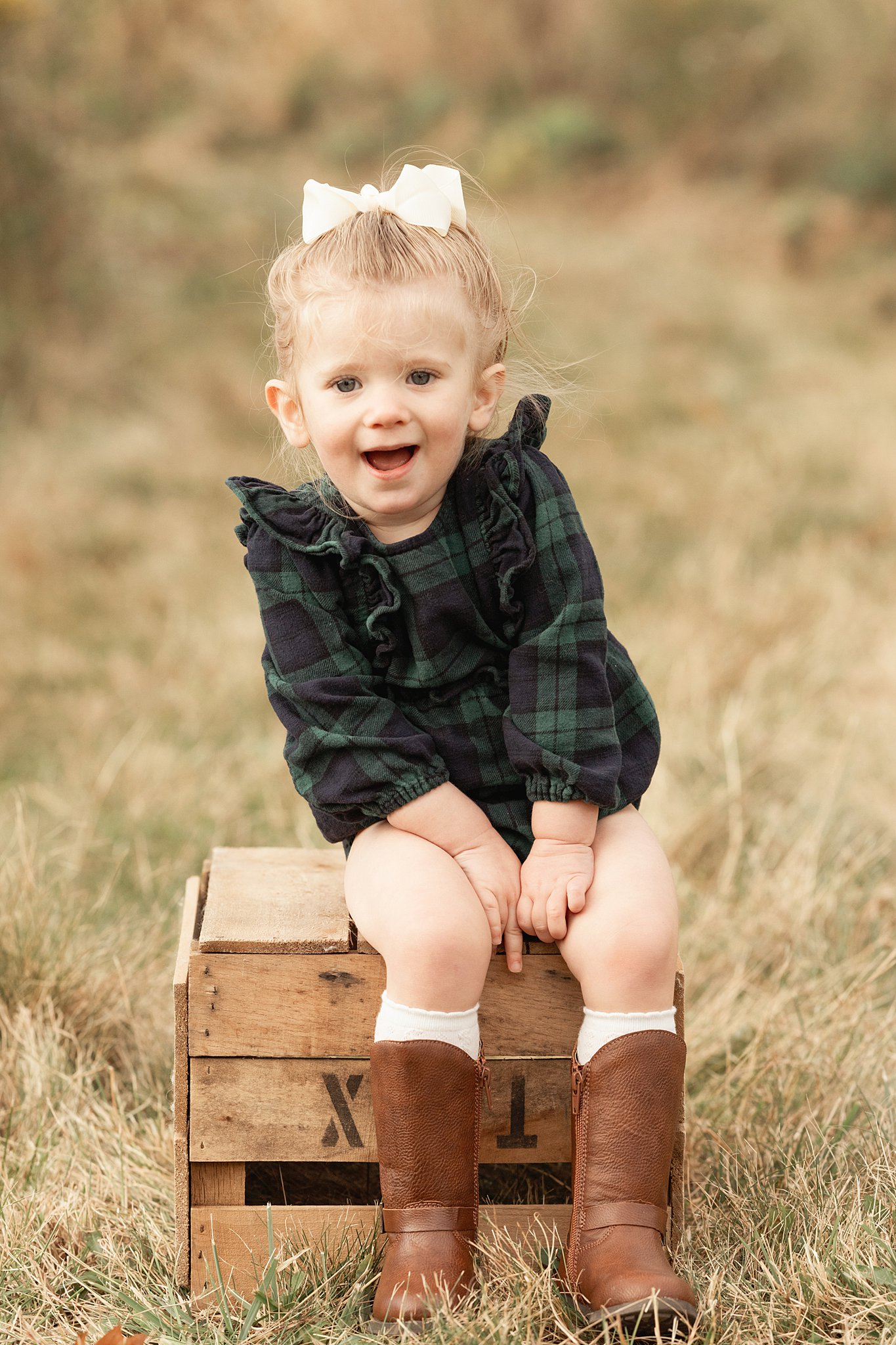 A young girl in a green plaid dress sits on a wooden box in a field after attending Pittsburgh Preschools