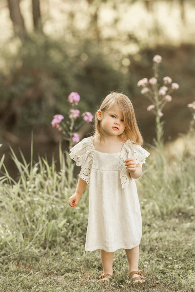 A toddler girl in a white dress stands in a field by wildflowers before visiting Pittsburgh Toy Stores