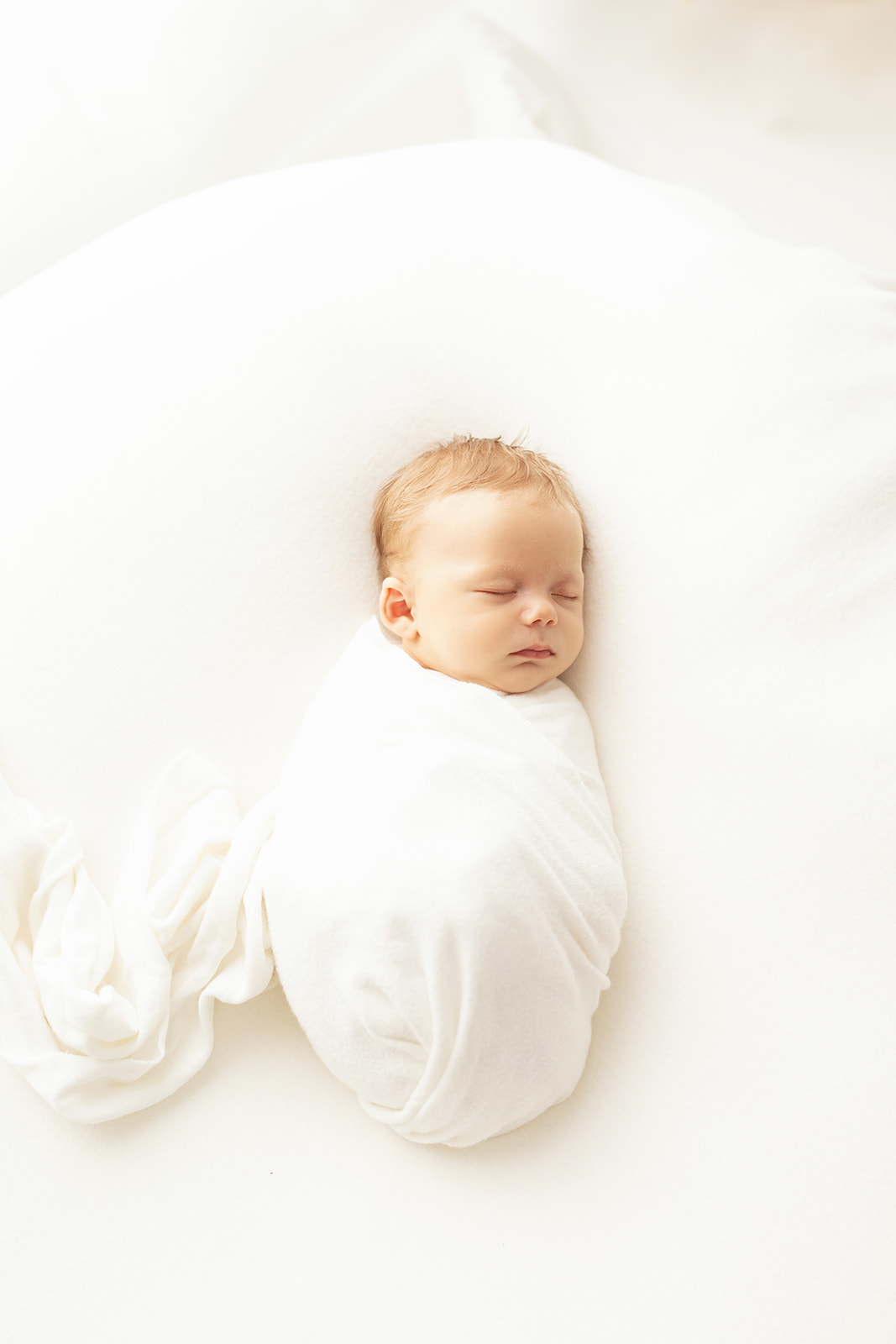A newborn baby sleeps in a white swaddle in a studio