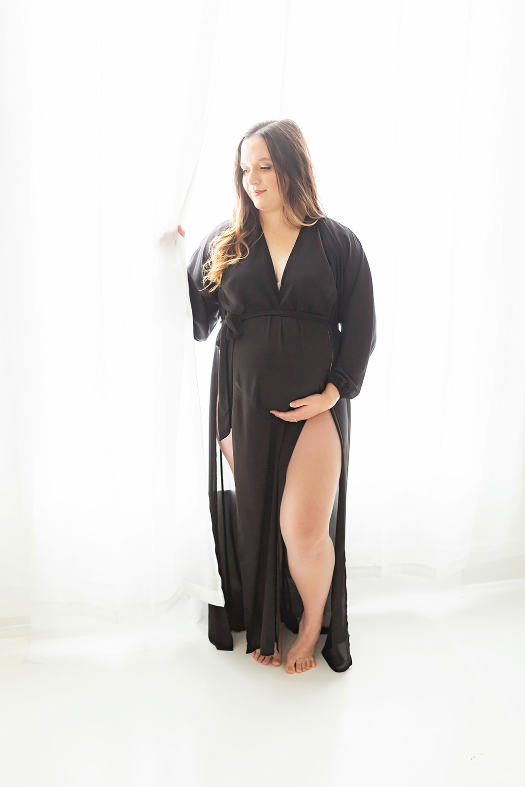 A mom to be stands in a window in a studio wearing a black maternity dress