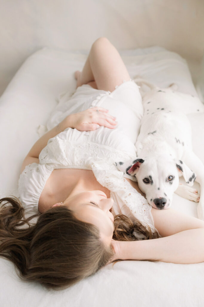expecting mom in white dress laying on white bed with dog next to her photographed by pittsburgh maternity photographer petite magnolia photography