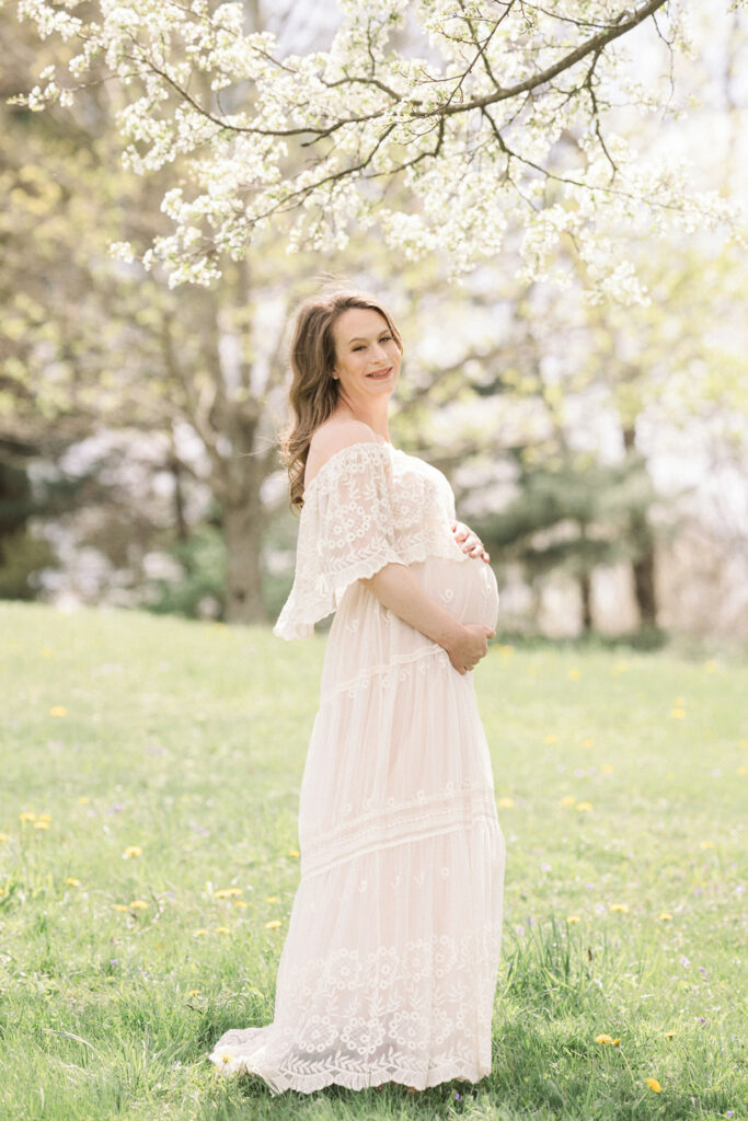 A happy mom to be in a white maternity gown holds her bump while standing in a grassy field