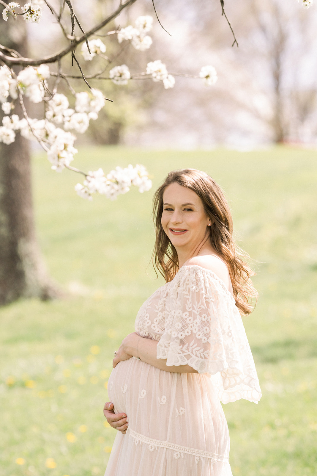 A mom to be smiles and holds the bump while standing in a white maternity gown under a blooming tree at sunset for Maternity Photos Pittsburgh