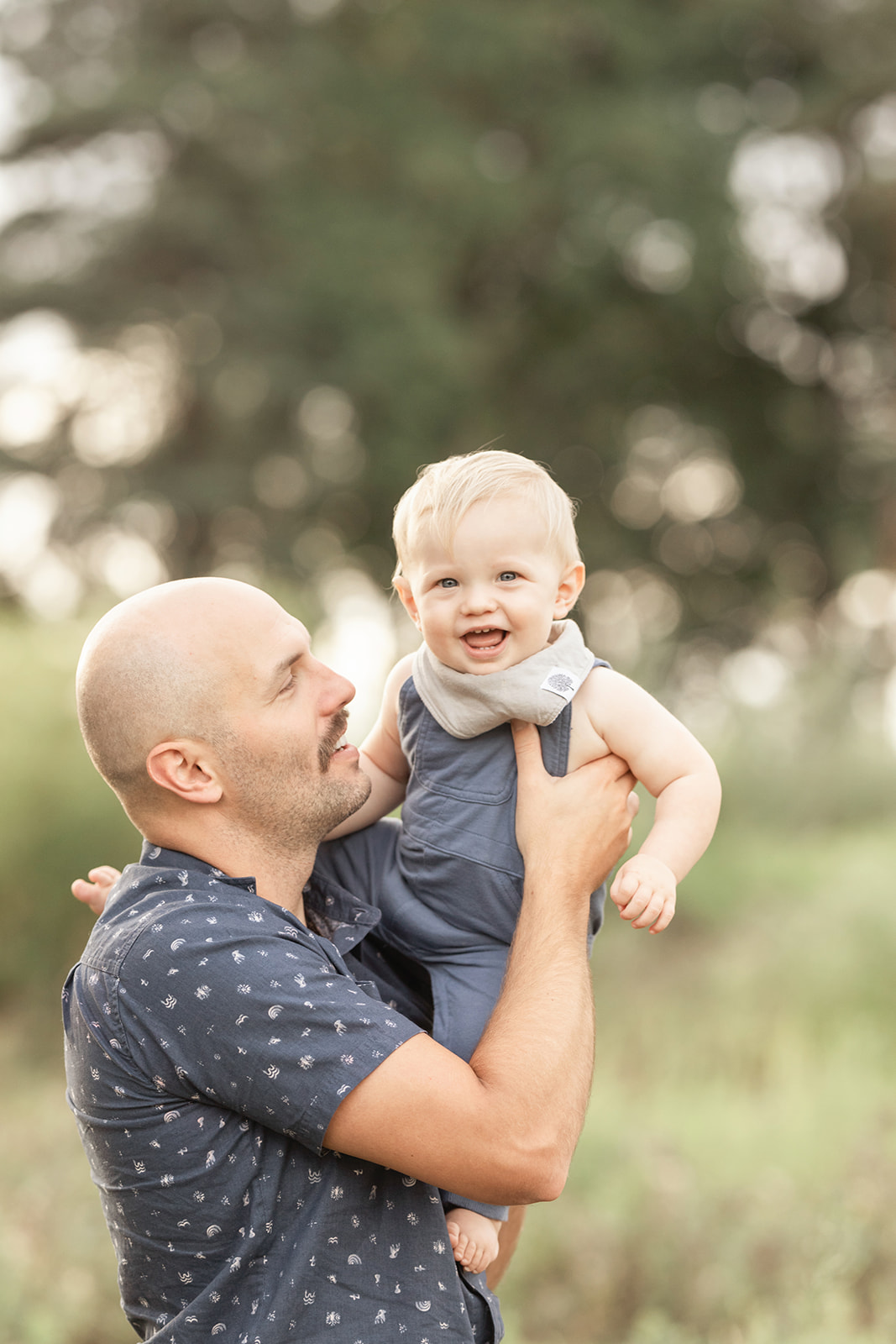 A father in a blue shirt lifts his laughing toddler son in blue overalls in a park at sunset