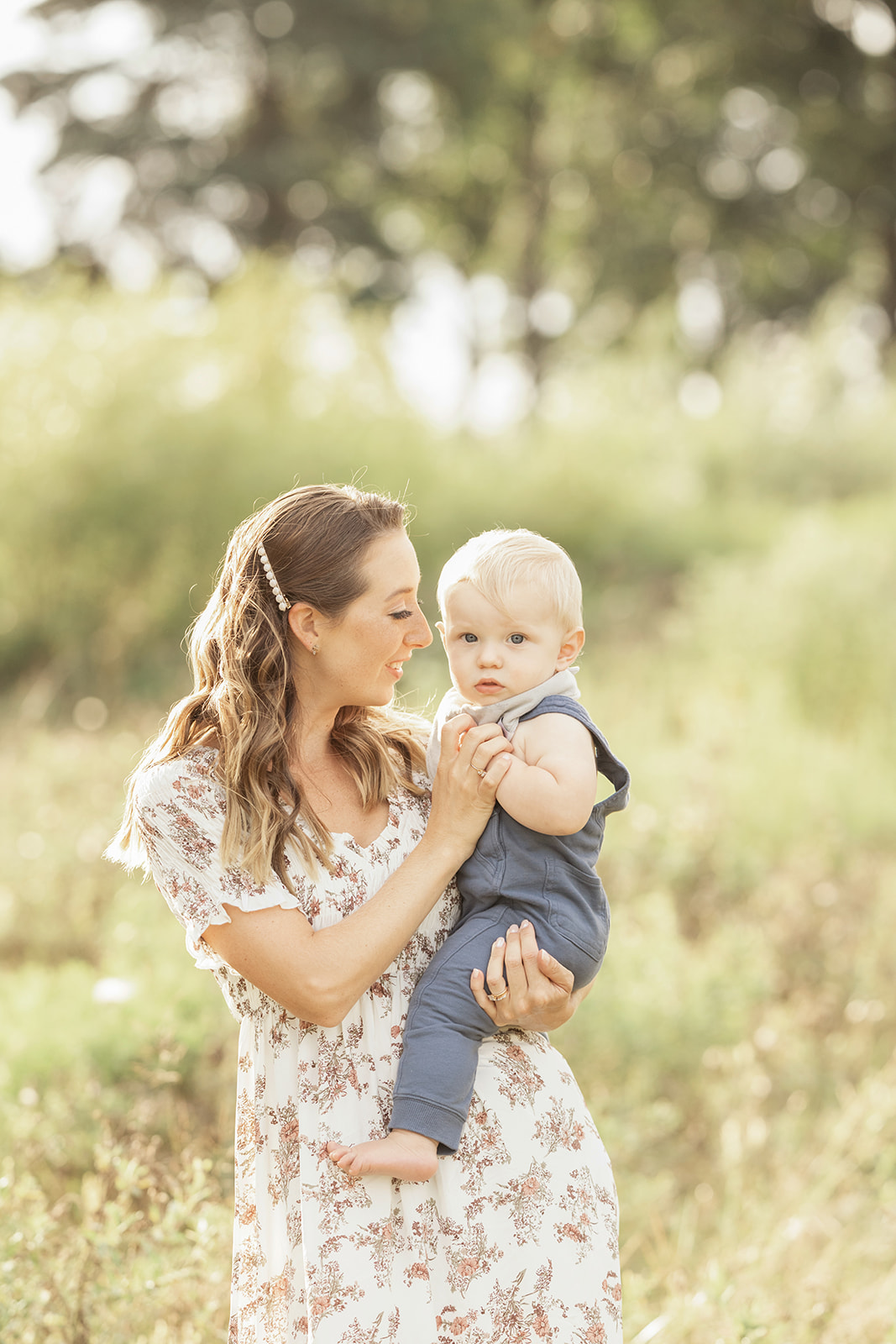 A happy mother in a floral dress plays with her toddler son on her hip while walking through a park at sunset