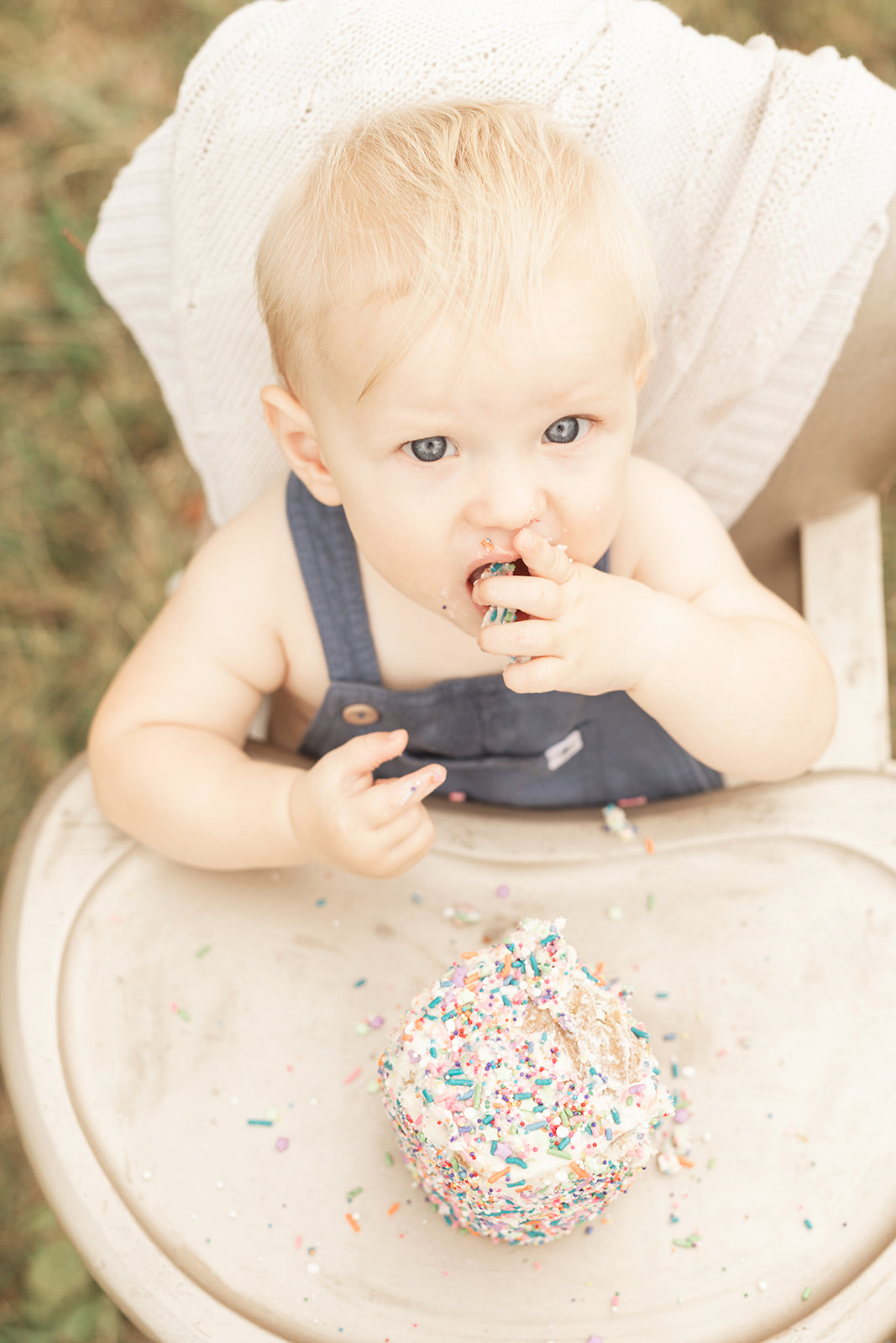 A toddler boy in blue overalls sits in a gold high chair in a park eating sprinkle covered cake for some Milestone Photography Pittsburgh