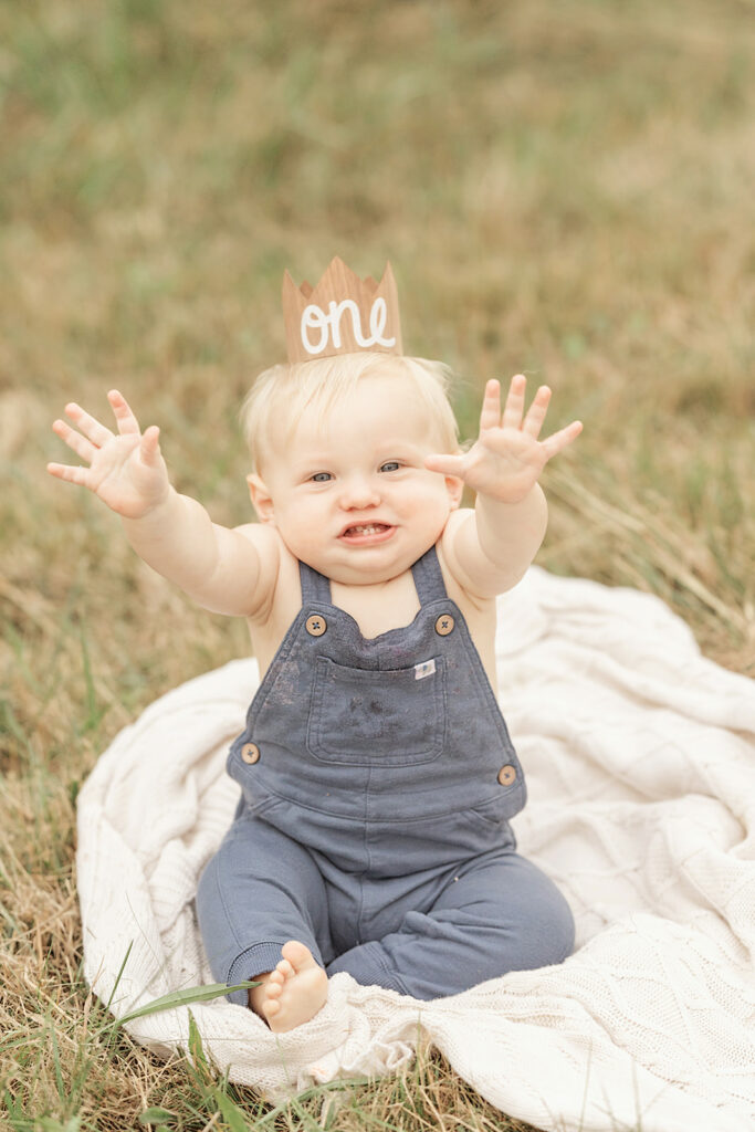 A toddler boy in blue overalls raising his hands while sitting in a park lawn for some Milestone Photography Pittsburgh