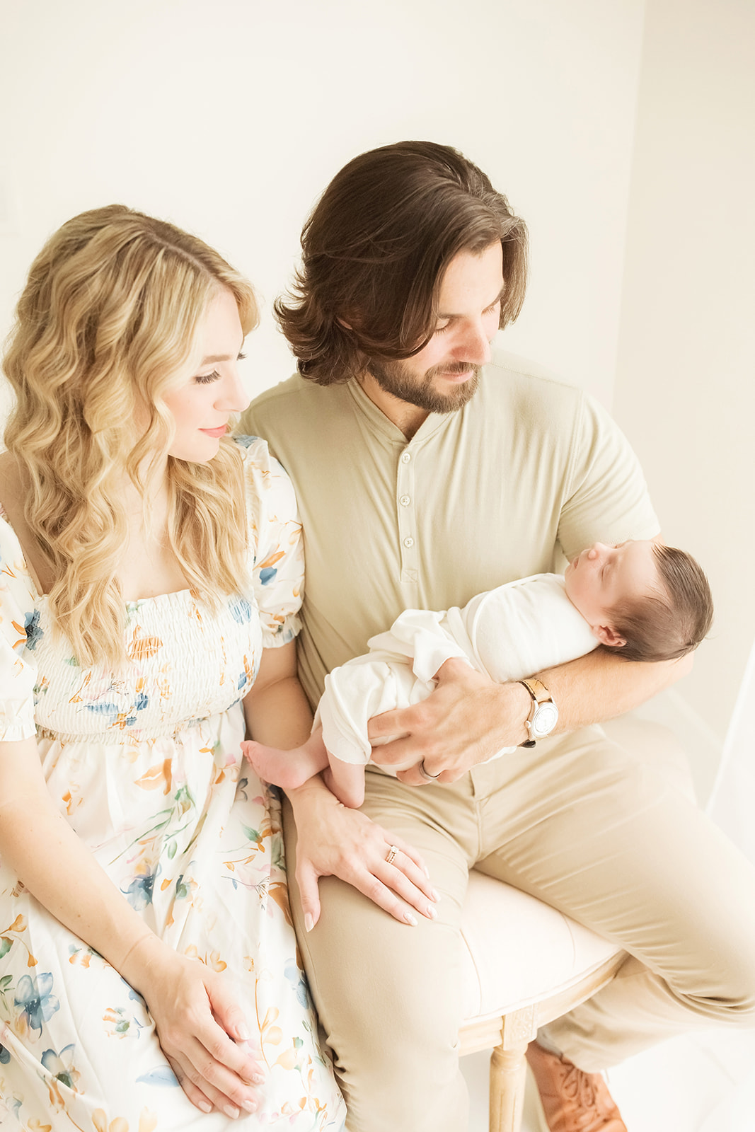 Happy new parents sit on a bench in a window holding their sleeping newborn baby in dad's arms