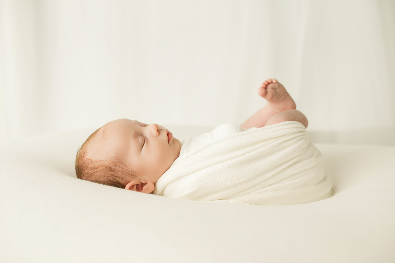 A newborn baby sleeps on a white pad while wrapped in a white swaddle