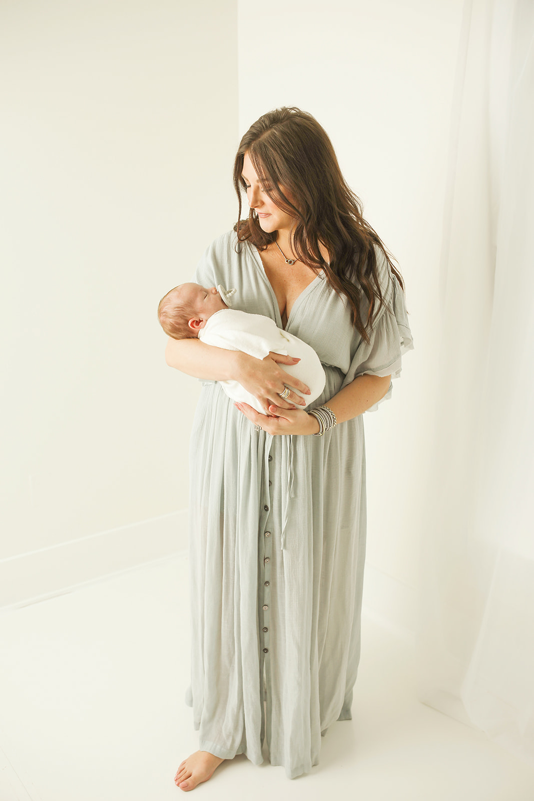 A new mother stands in a studio in a green dress smiles down at her sleeping newborn baby in her arms