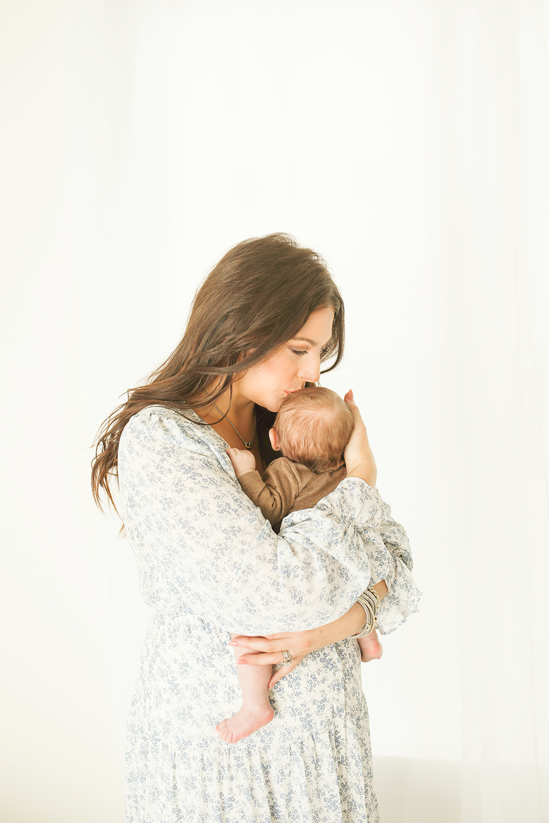 A happy new mother in a white floral dress kisses her newborn baby in her arms while standing in a studio for a Pittsburgh Photographer