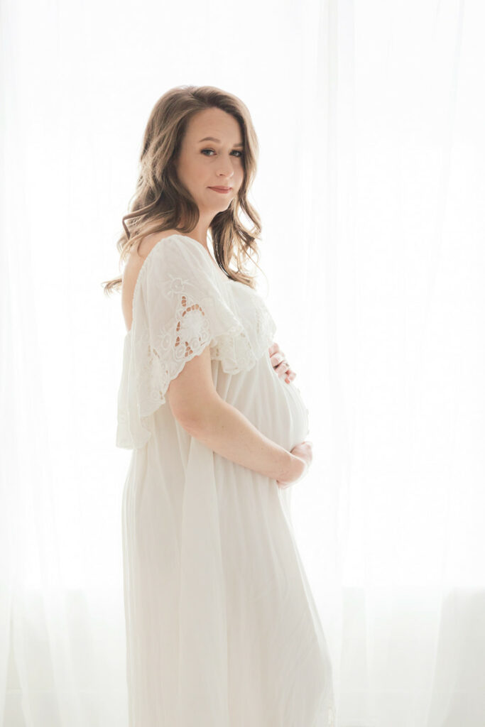 A happy mother to be stands in a studio window in a white maternity gown with lace collar holding her bump