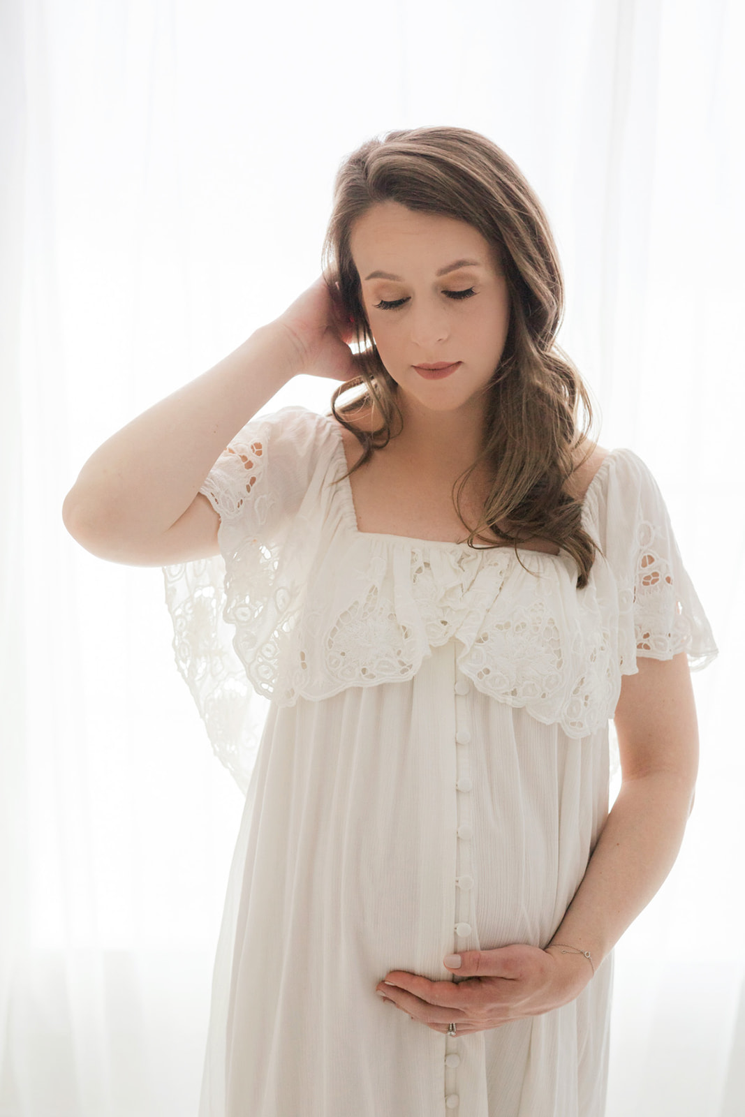 A mom to be in a white maternity gown stands in a studio window with a hand in her hair holding her bump after visiting Birthright Of Pittsburgh