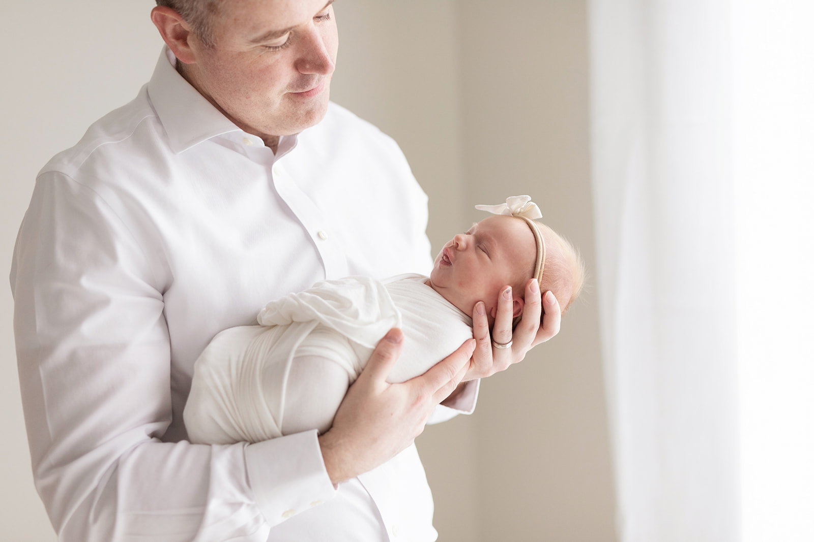 A newborn baby sleeps in dad's hands as he stands in a window with tongue out