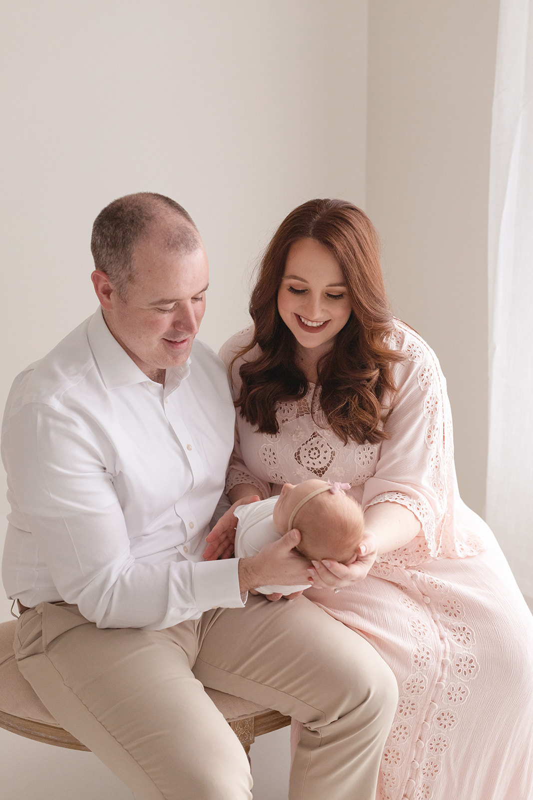 Happy mom and dad sit in a studio on a bench in a window smiling down at their newborn daughter in their laps before meeting an Au Pair Pittsburgh