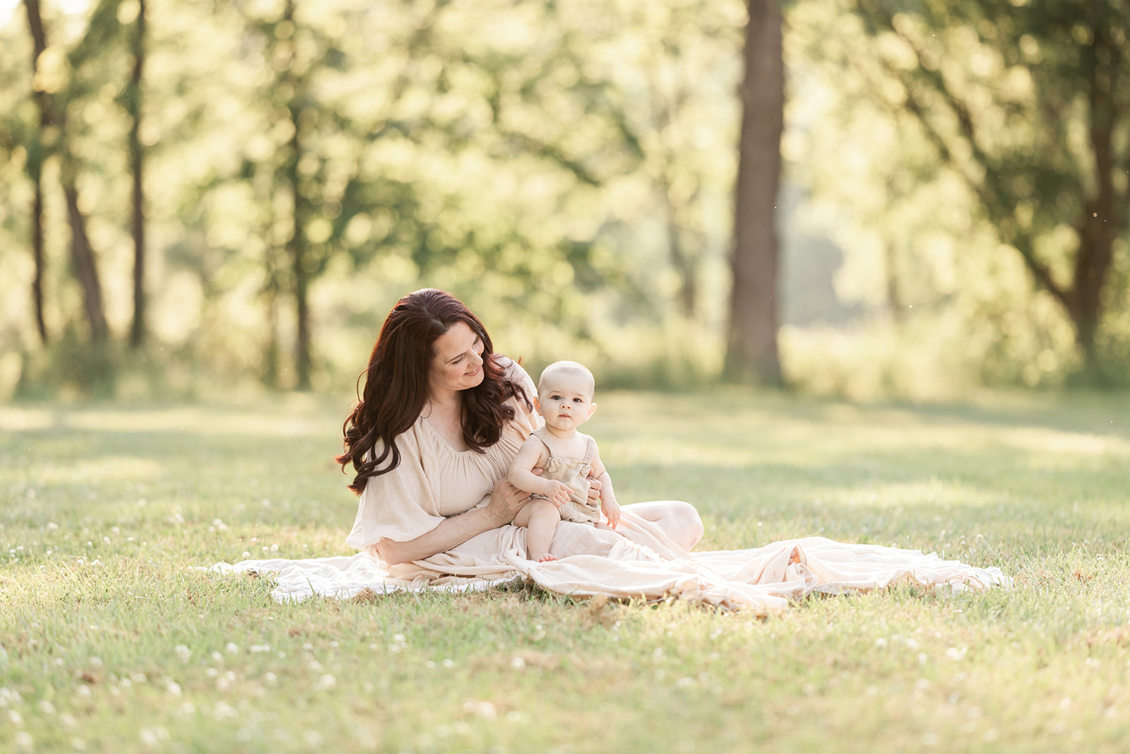 A happy mom sits in a park lawn while her infant sits in her lap in tan overalls