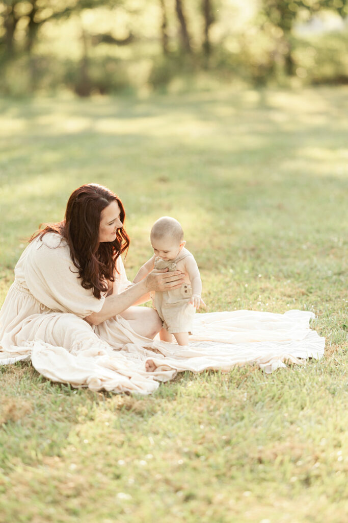 A mom sits on a picnic blanket in a park lawn in a white dress playing with her infant child after some baby music classes Pittsburgh