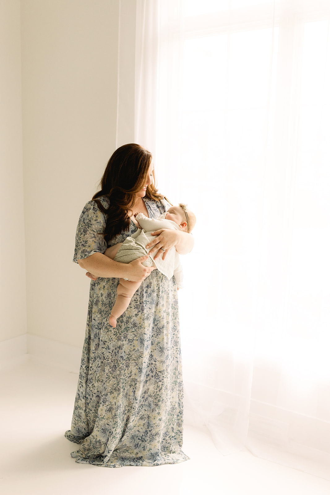 A happy mom smiles while cradling her sleeping infant in her arms while standing in a window thanks to Diaper Service Pittsburgh