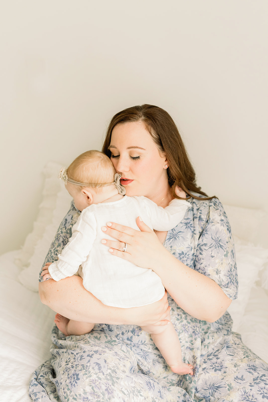 A mother sits on a bed in a blue print dress playing with her infant daughter in her lap while using Diaper Service Pittsburgh