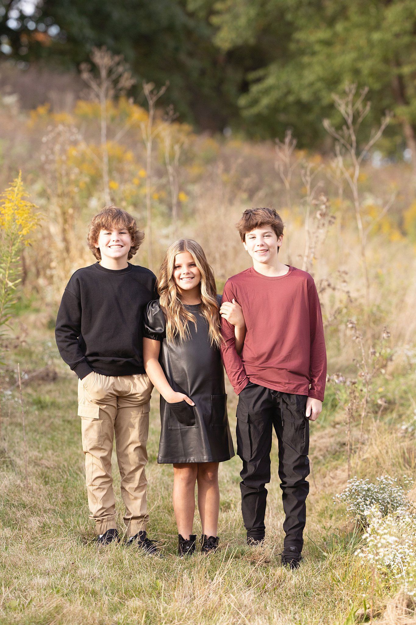 A young girl holds onto the arms of her older brother while standing with her other older brother in a field of tall grasses