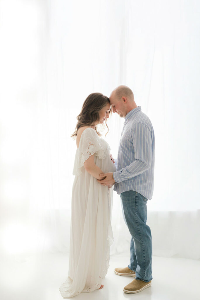 Expecting parents stand touching foreheads and holding the bump together in a large studio window