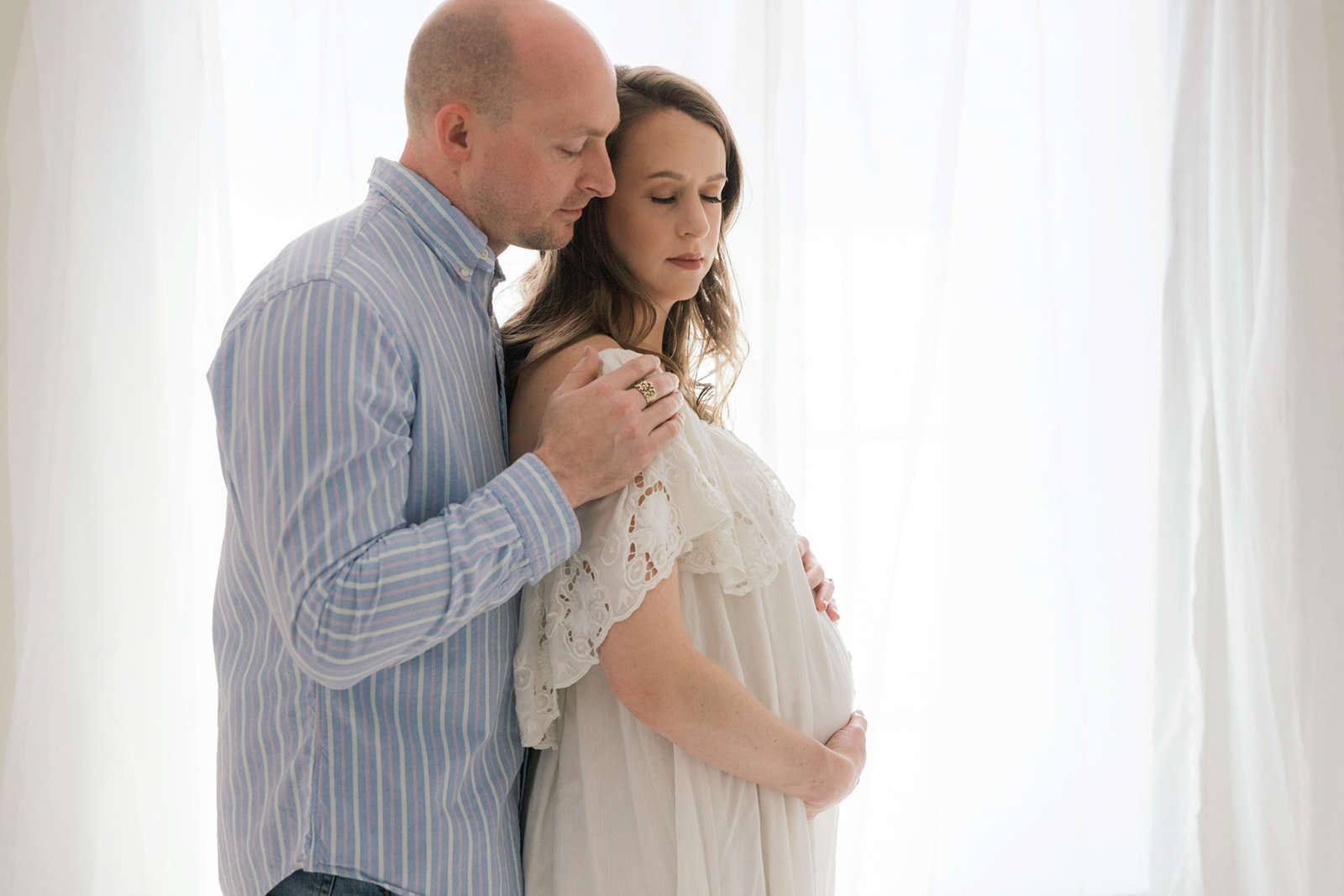 A man in a stripe shirt embraces his pregnant wife as they stand in a window after meeting Pittsburgh Doula Network