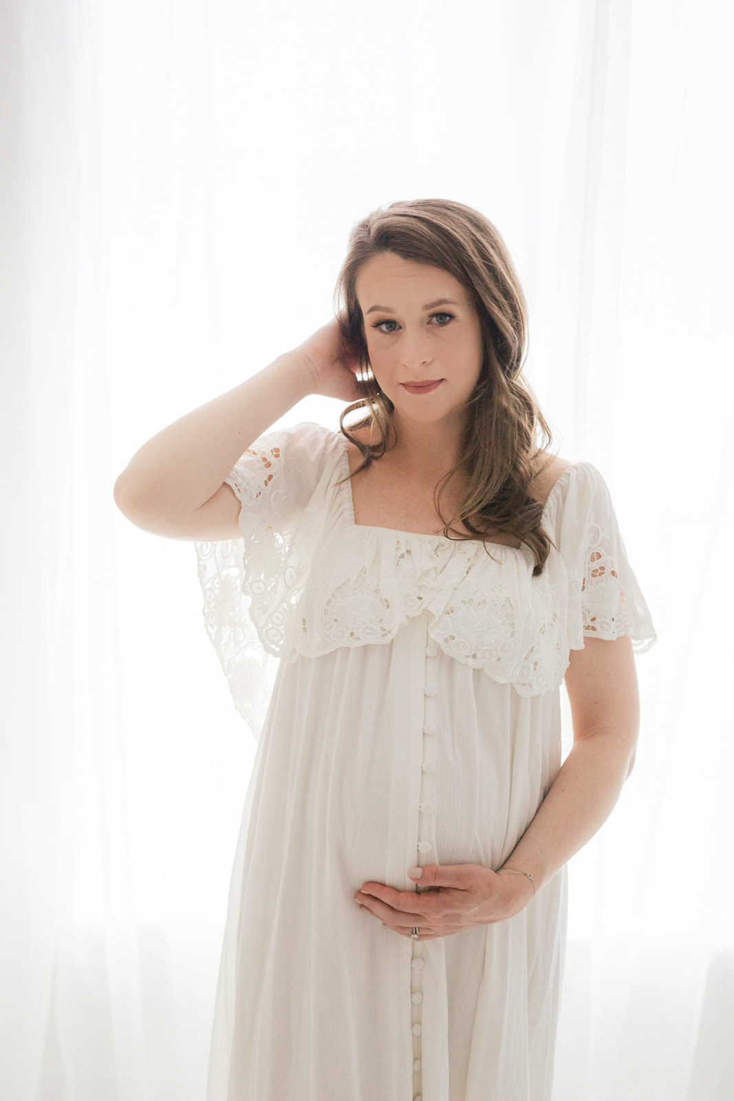 A mom to be in a white dress stands in a window holding her bump and hair back after visiting Pittsburgh Doula Network