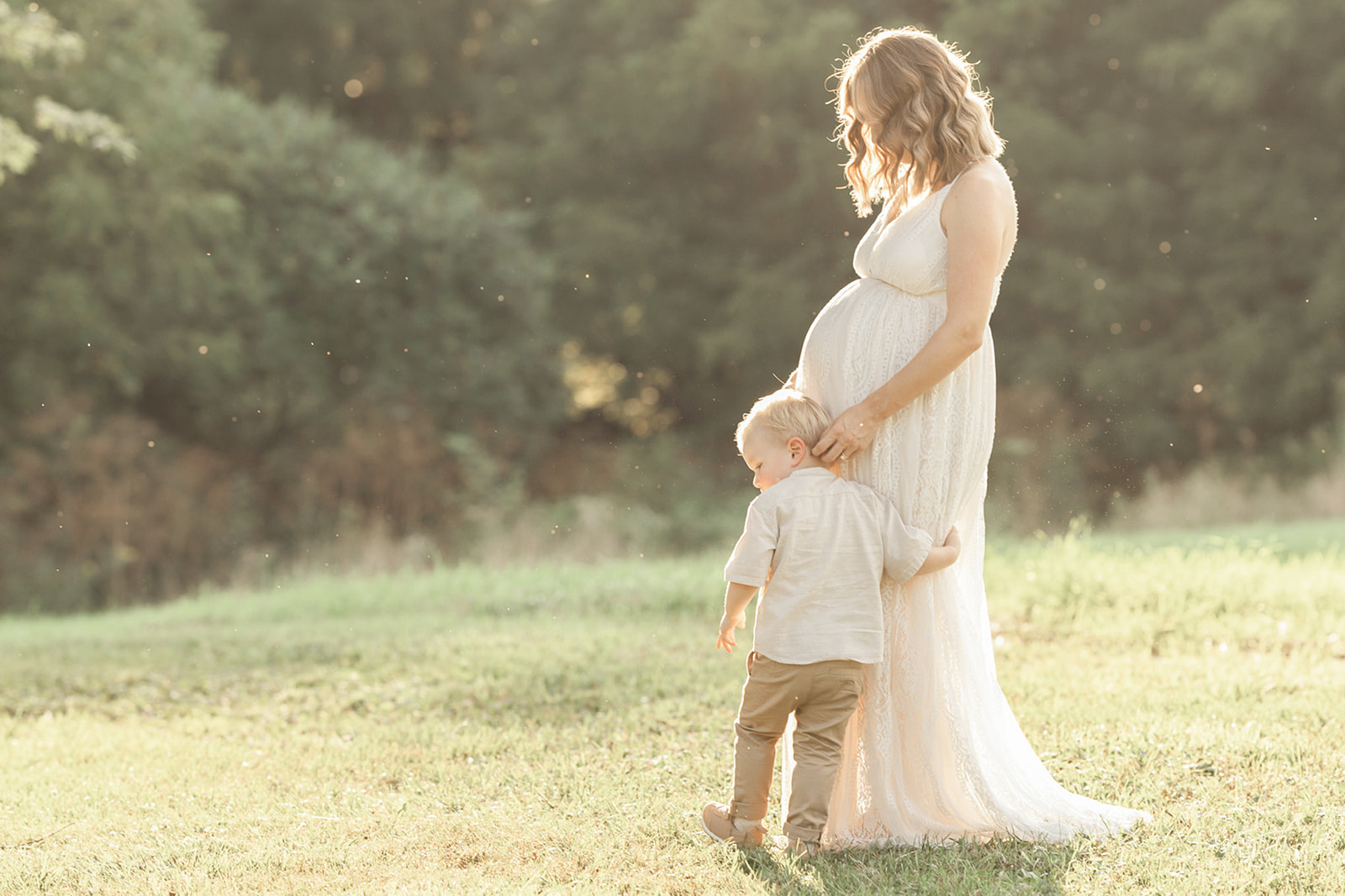 A pregnant mother in a white lace maternity gown explores a field at sunset with her toddler son