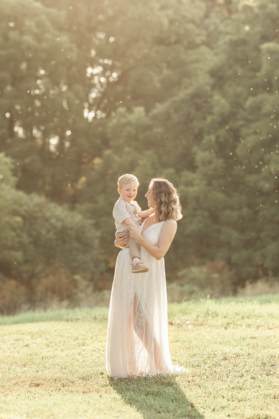 A pregnant mother in a lace maternity gown stands in a field at sunset with her toddler sitting on the bump thanks to UPMC OBGYN