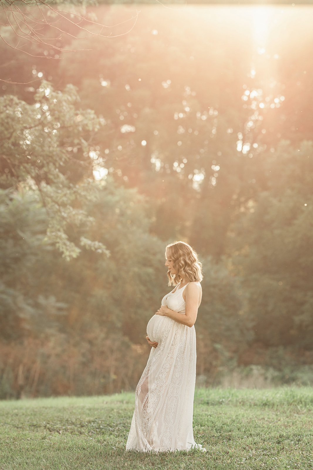 A mom to be in a long white maternity gown stands in a lawn at sunset after some Fertility Acupuncture Pittsburgh