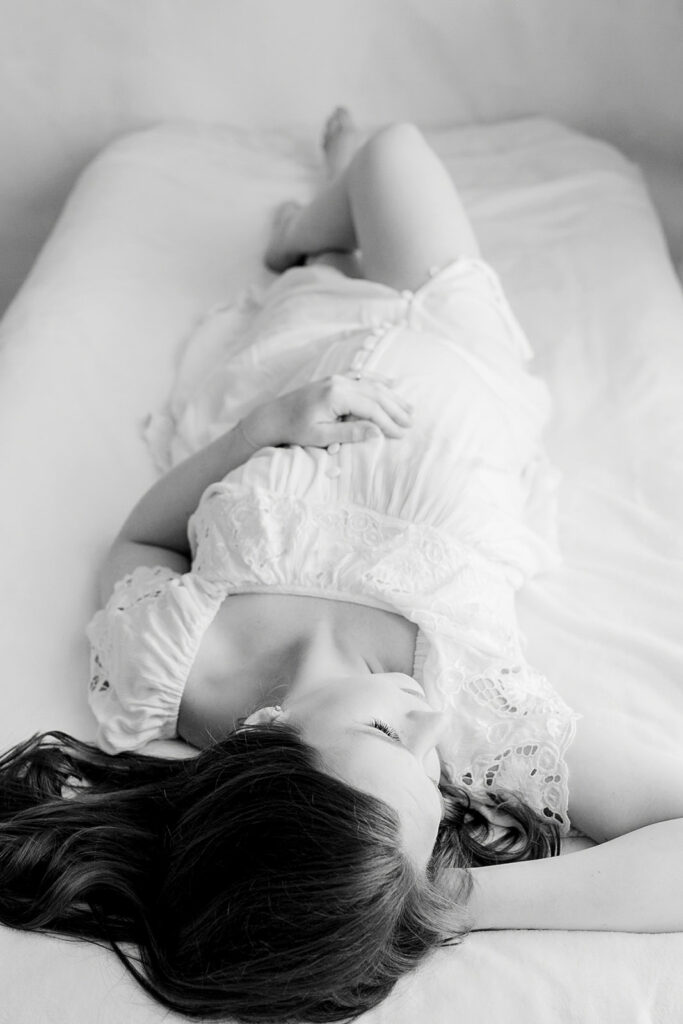 A pregnant woman lays on a bed in a white maternity gown