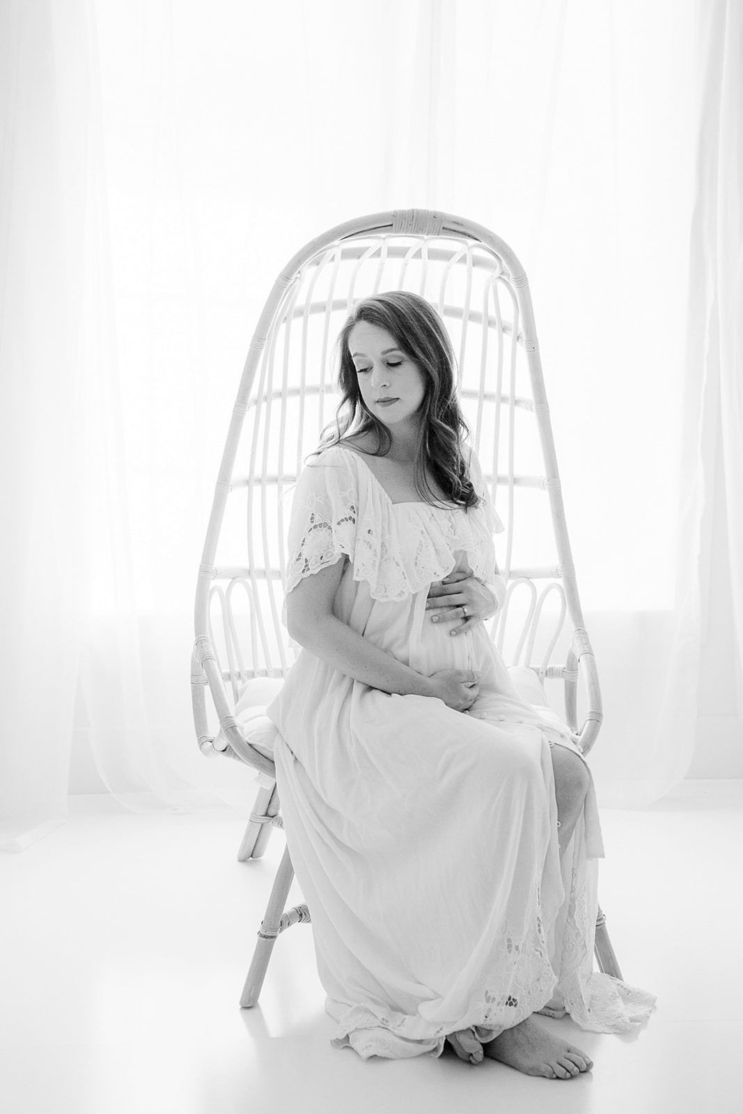 A mom to be sits in a wicker chair in a studio window holding her bump in a long white maternity gown before some Pelvic Floor Therapy Pittsburgh