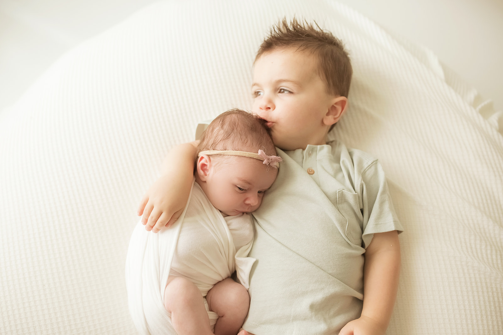 A newborn baby girl cuddles on her bigger brother while laying on a bed in a studio