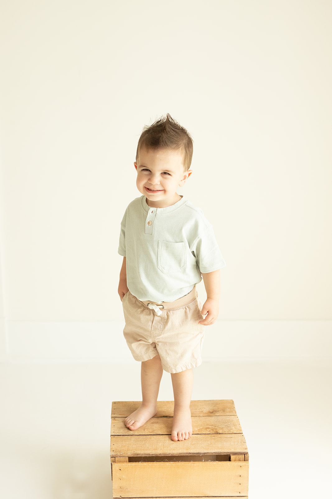 A smiling toddler boy stands on a wooden crate in a studio smiling after meeting Pittsburgh Babysitters