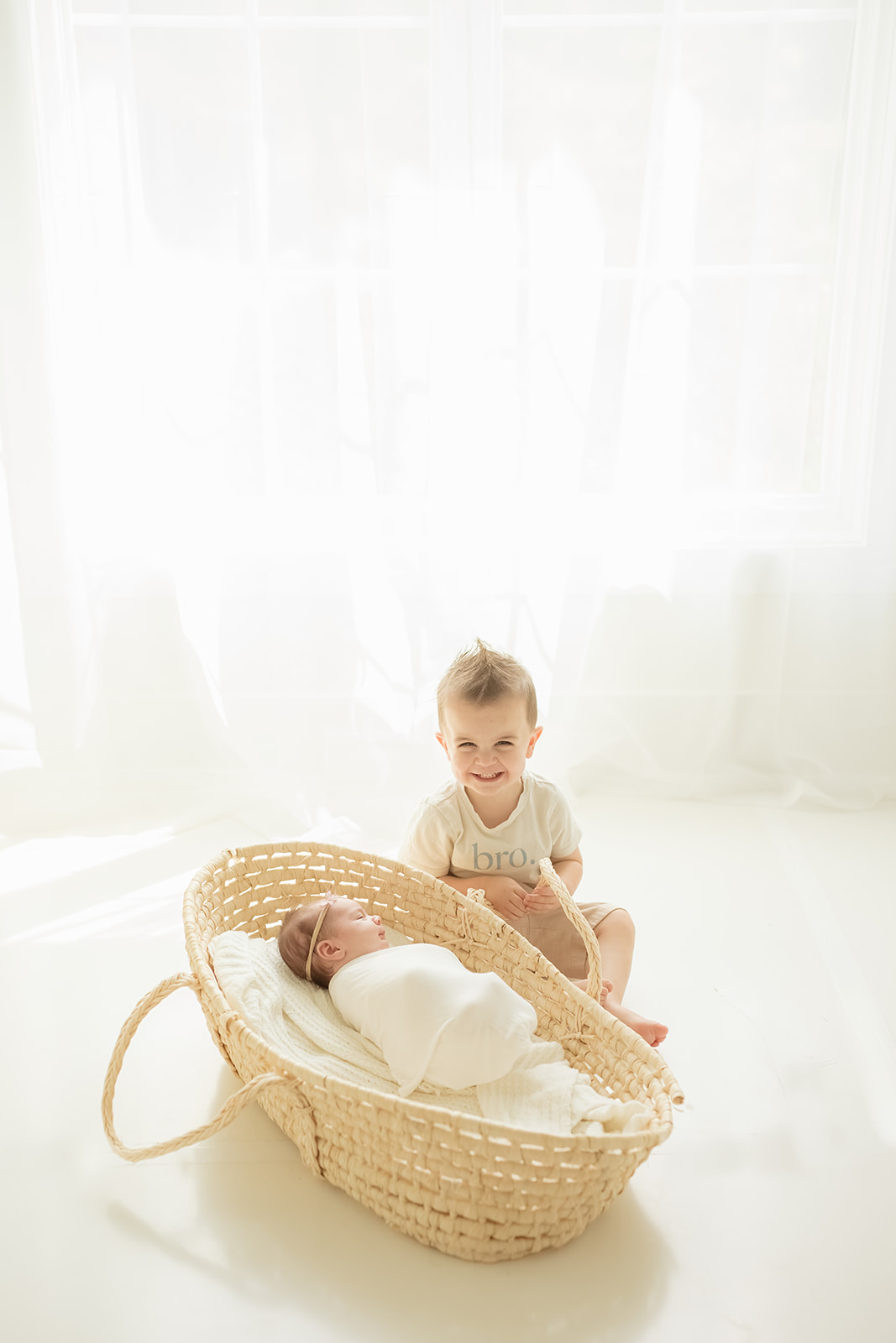 A toddler boy sits on the floor under a window with his newborn baby sister sleeping in a woven basket after meeting Pittsburgh Babysitters