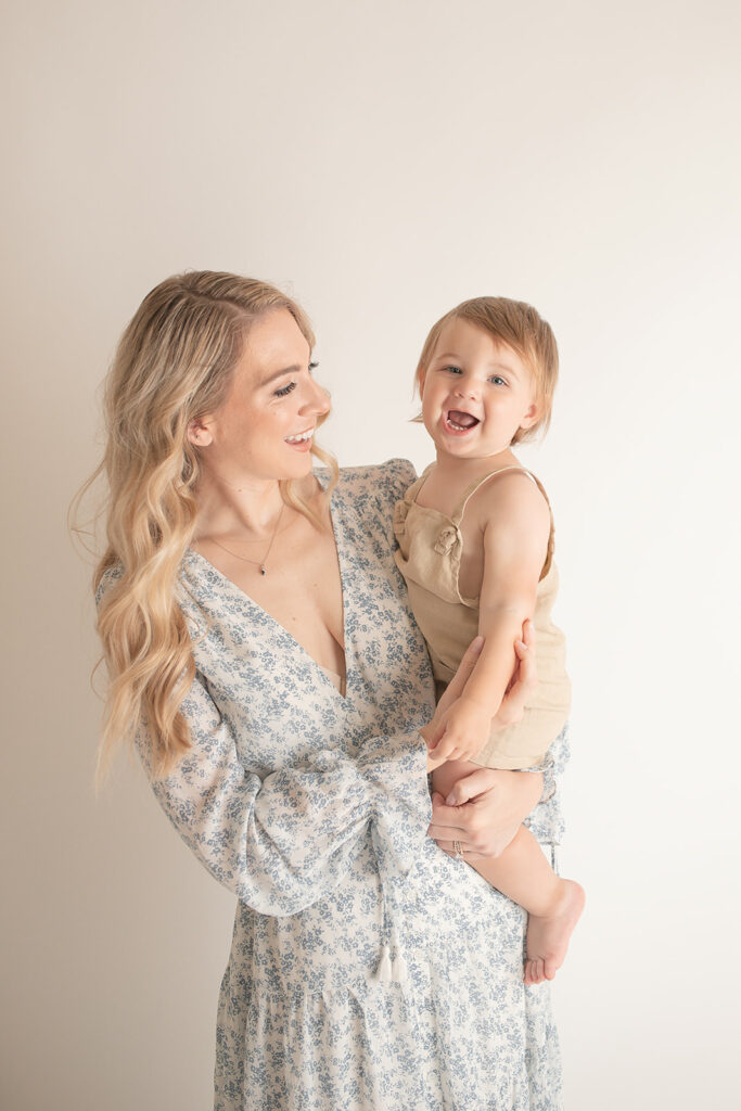 A laughing toddler sits on mom's hip while she smiles along in a studio