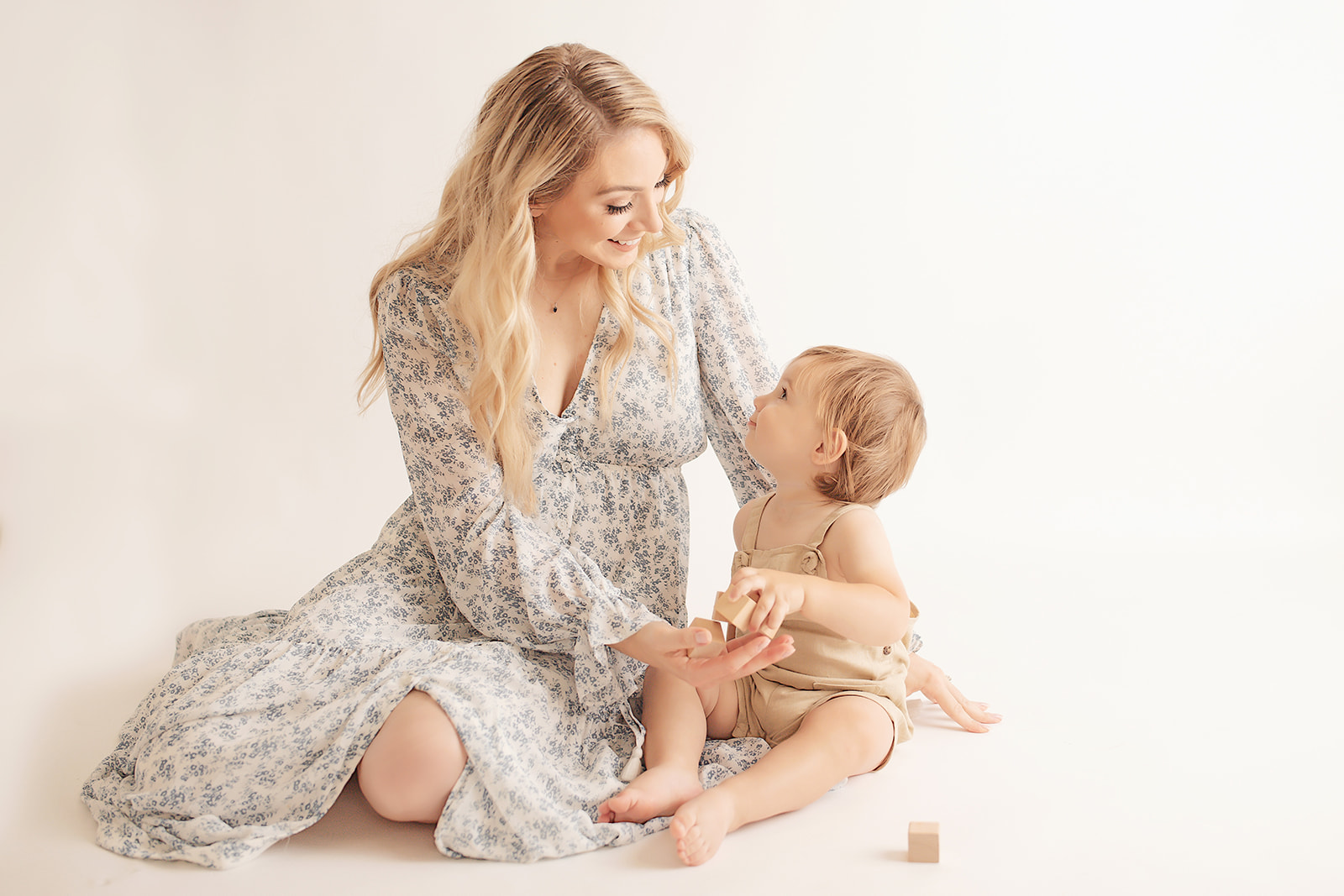 A happy mother sits on the floor of a studio in a blue dress playing with her toddler son and wood blocks before some Pittsburgh birthday parties for kids