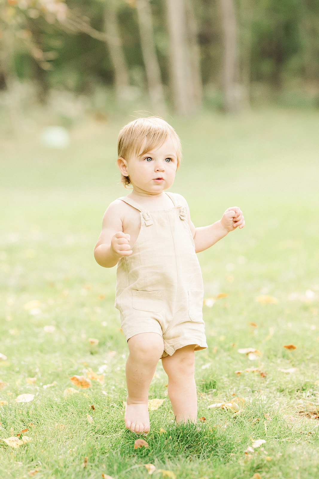 A toddler boy in tan overalls explores a park lawn barefoot after visiting Pittsburgh Montessori Schools