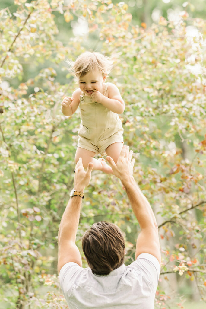 A father tosses his happy toddler up in the air in a park after exploring Pittsburgh Montessori Schools