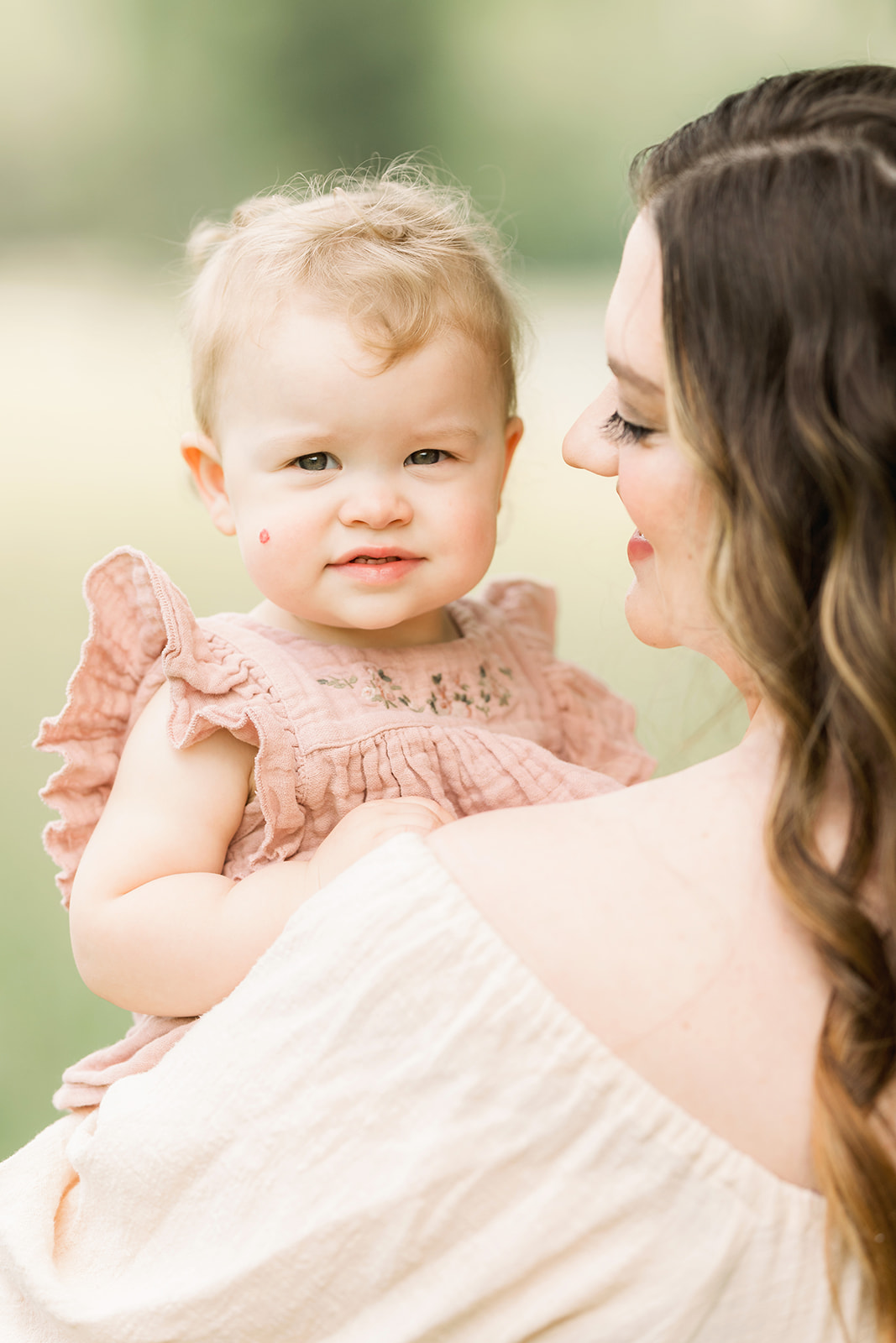 A toddler girl smiles while sitting in mom's arms in a field in a pink dress