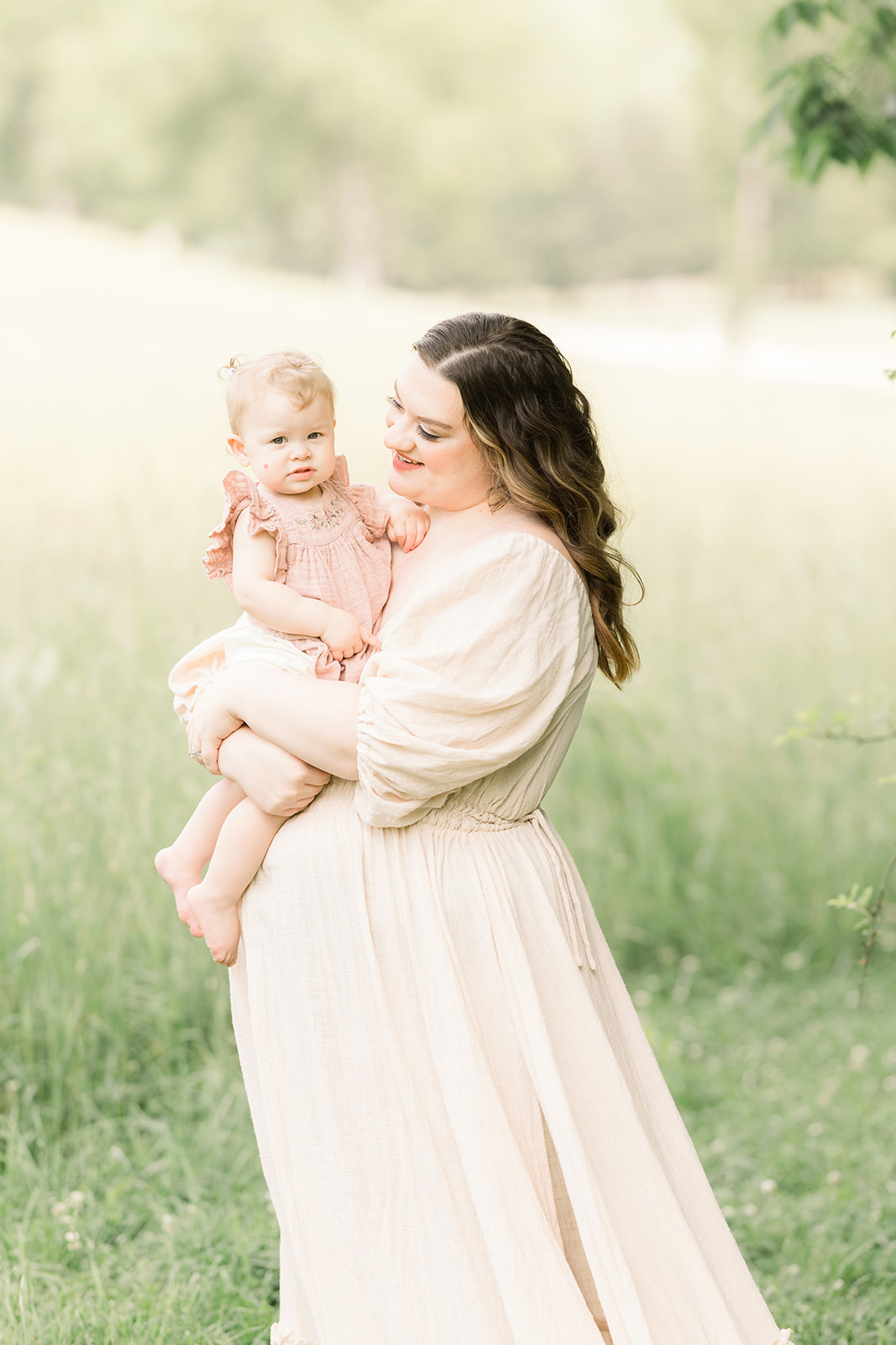 A happy mom smiles at her toddler daughter in her arms in a pink dress in a field after some Pittsburgh Parenting Classes