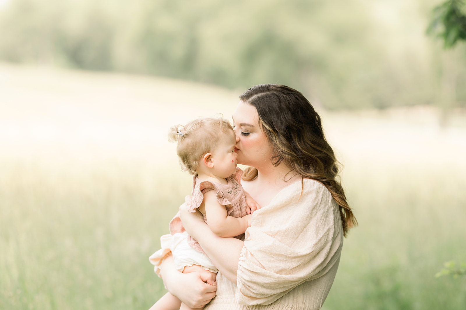 A mother in a cream dress kisses the cheek of her toddler in her arms in a field of grass after some Pittsburgh Parenting Classes